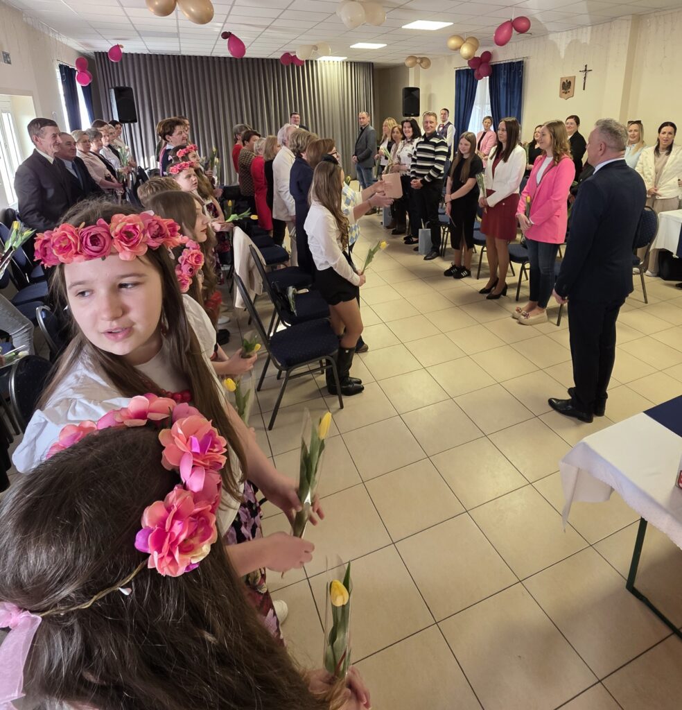 Grupa ludzi stoi w jasnym pomieszczeniu, dzieci z kwiatami, dekoracje balonowe. Tło w odcieniach beżu i niebieskie zasłony.