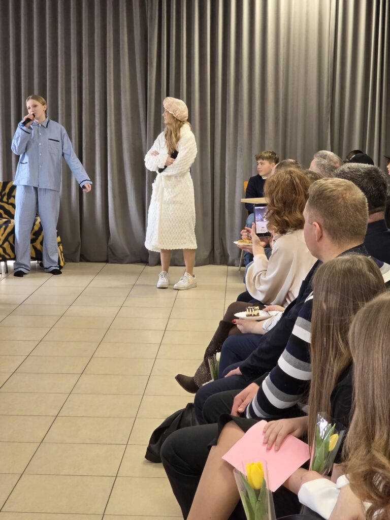 Na zdjęciu widać dwie osoby na scenie, jedna w jasnoniebieskim stroju, druga w białym szlafroku i czepku. W tle znajdują się szare zasłony, a publiczność siedzi z boku, trzymając żółte tulipany. Tło jest