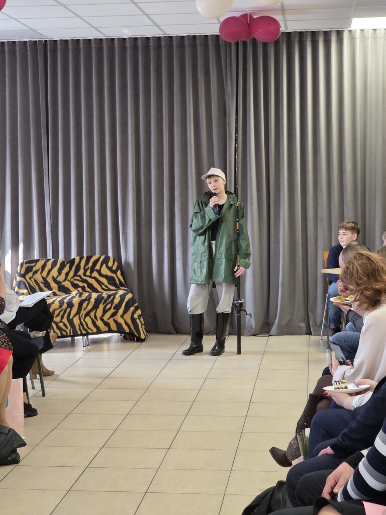 Na zdjęciu występuje osoba w zielonej kurtce i białej czapce, trzymająca mikrofon. W tle są szare zasłony i sofa z żółto-czarnym pokrowcem. Wokół siedzą ludzie na krzesłach.