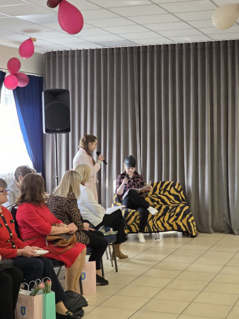 Sala z szarymi zasłonami i różowymi balonami. Ludzie siedzą, dwie osoby brały udział w prezentacji z mikrofonami. Tło to zasłony i kanapa w tygrysi wzór.