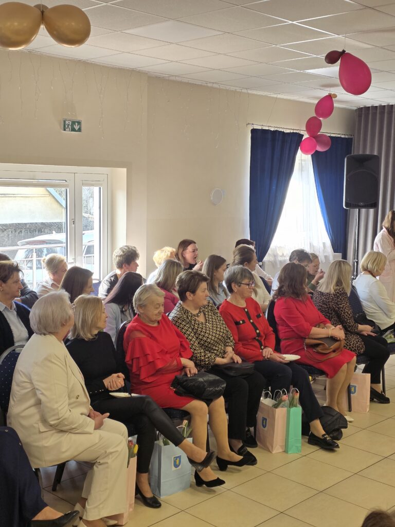 Grupa osób siedzi w sali z balonami na suficie. Większość ubrana jest w czerwone i czarne stroje. W tle niebieskie zasłony i jasne ściany.