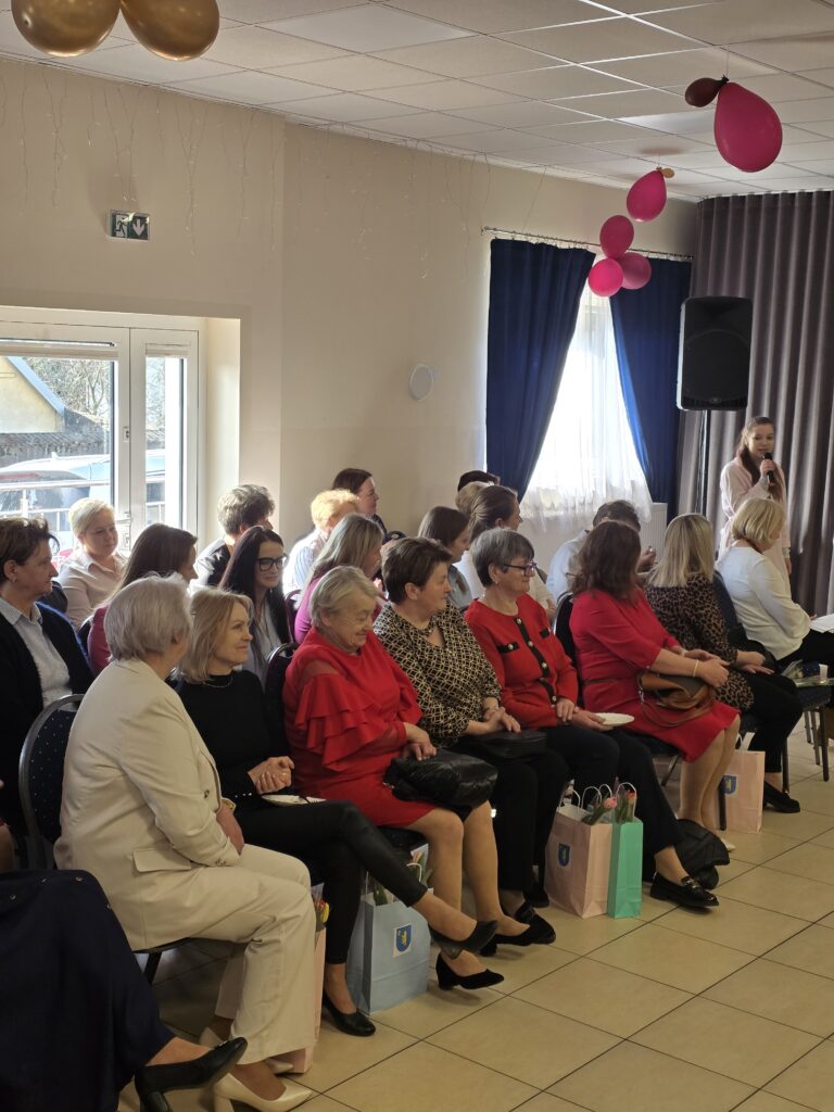 Grupa osób siedzi na krzesłach w jasnym pomieszczeniu, z oknami z przodu. Na suficie wiszą różowe i złote balony. W tle stoi osoba przemawiająca przy mikrofonie.