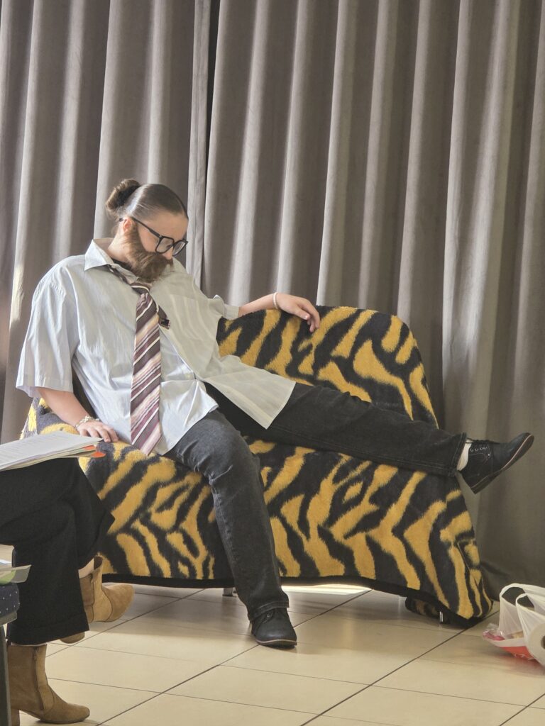 Osoba z brodą i okularami siedzi na sofie z wzorem tygrysim w żółto-czarne paski. Ubrana jest w koszulę, krawat i ciemne spodnie. W tle szare zasłony, a obok na podłodze leżą przed