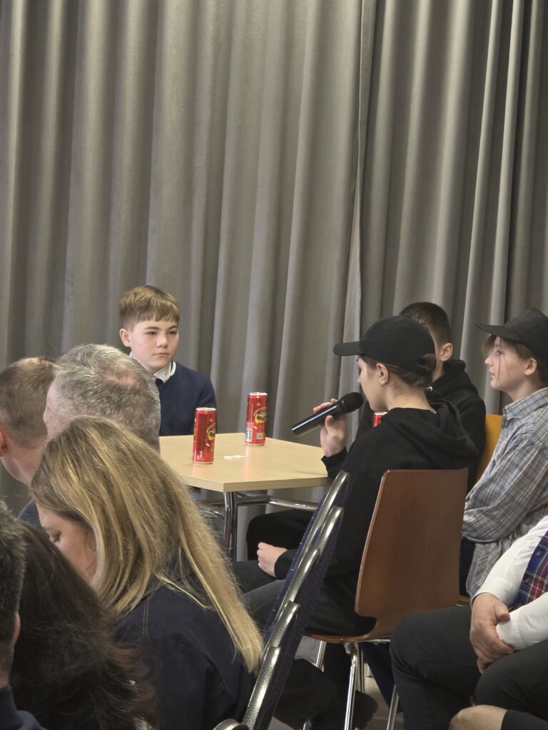 Młodzież siedzi przy stole, rozmawiając. Jedna osoba trzyma mikrofon. Na stole stoją czerwone puszki napoju. W tle szara zasłona.