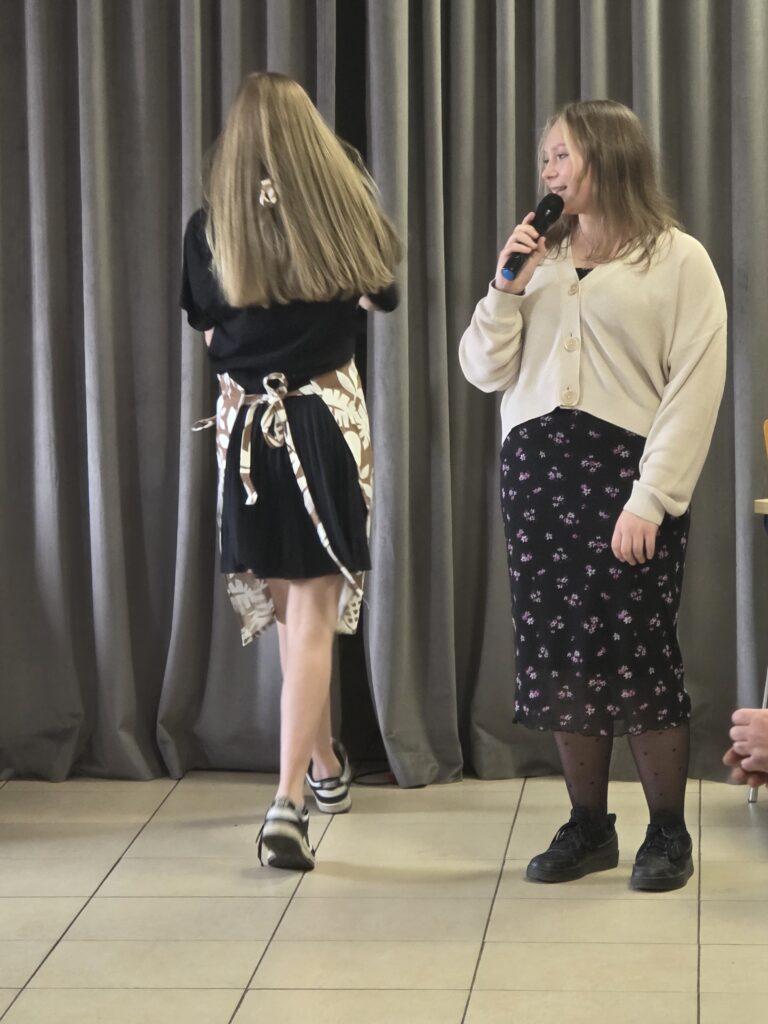 Na zdjęciu są dwie dziewczyny, jedna trzyma mikrofon. Tło stanowią szare zasłony. Dziewczyna po lewej ma jasne, rozpuszczone włosy.