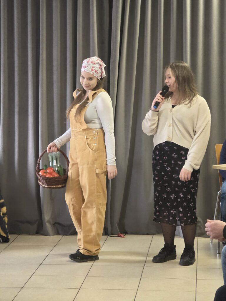 Dwie kobiety stoją na tle szarej zasłony. Jedna trzyma kosz z warzywami, ubrana w jasny strój roboczy. Druga mówi do mikrofonu.