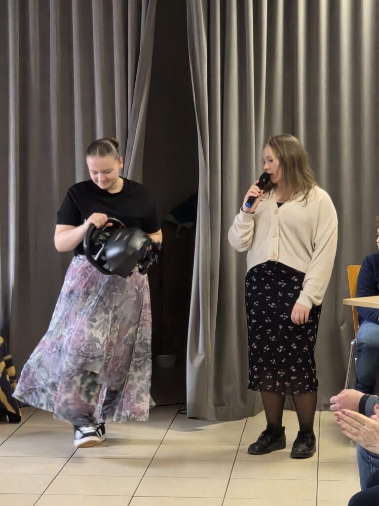 Dwie kobiety stoją na tle szarych zasłon, jedna trzyma kask, druga mówi do mikrofonu. Tło jest jasne, podłoga kremowa. Kobiety są ubrane w spódnice z kwiatowymi wzorami.