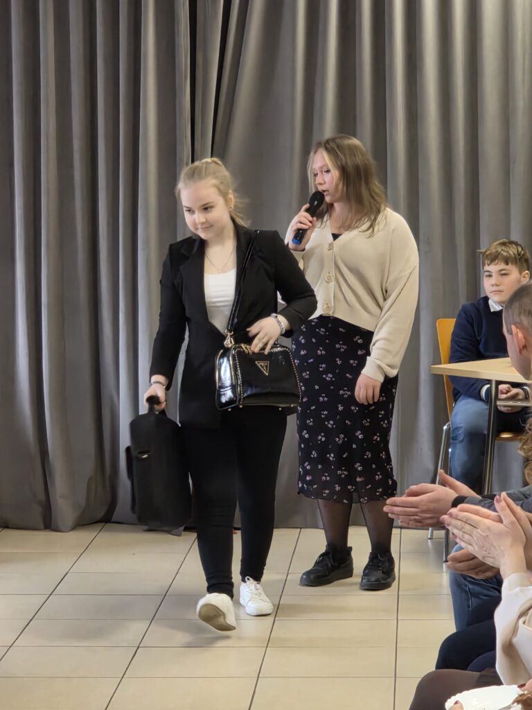 Młoda kobieta w czerni przechodzi obok publiczności; za nią osoba z mikrofonem. Szare zasłony w tle. Klaskający widzowie.