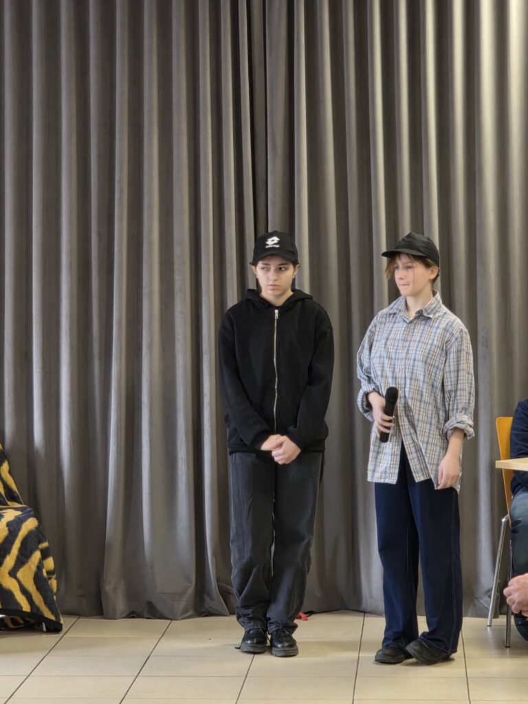 Na zdjęciu dwie osoby stoją na tle szarej zasłony. Jedna osoba trzyma mikrofon i ma koszulę w kratę, druga jest w czarnym ubraniu. Widoczny jest również skrajnie fragment krzesła po prawej stronie.