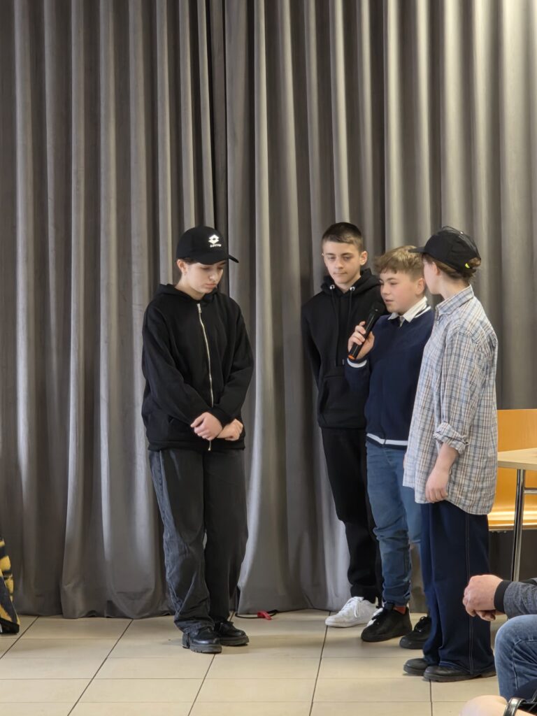 Grupa czterech chłopców stoi na tle szarych zasłon. Jeden z nich trzyma mikrofon, ubrani na czarno i w casualowe ubrania. Tła dopełniają szare zasłony i jasna podłoga.