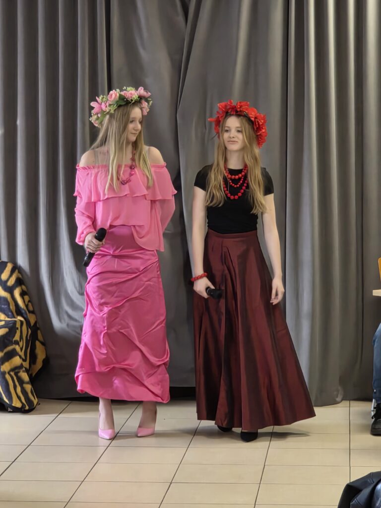 Dwie kobiety w kwiatach na głowach, ubrania w różu i czerwieni. Szare zasłony w tle, trzymają mikrofony. Podłoga wyłożona płytkami.