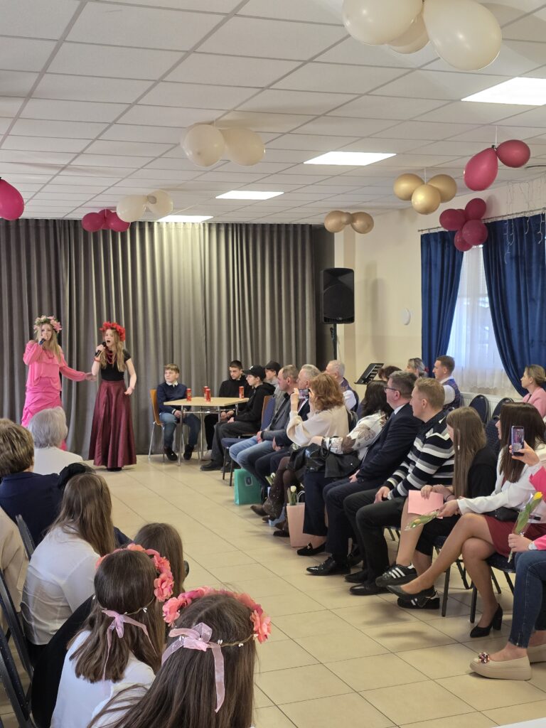 Na zdjęciu jest występ dwóch osób w kolorowych strojach w sali z publicznością. Tło stanowią szare zasłony i balony w ciepłych barwach. Widzowie siedzą w rzędach, niektórzy robią zdjęcia.