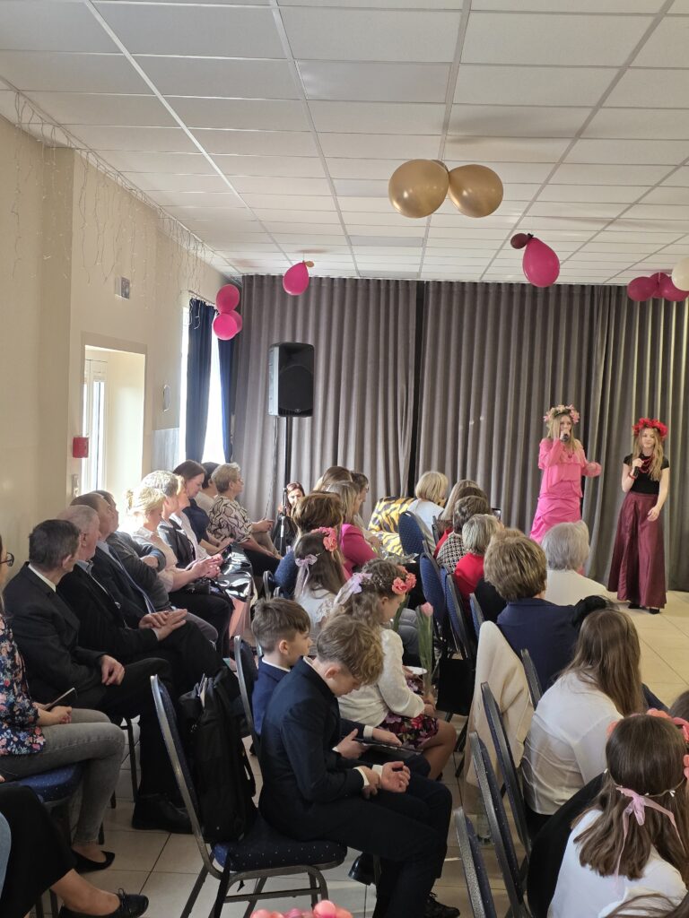 Na zdjęciu widać grupę osób siedzących w sali z szarymi zasłonami. Na suficie wiszą różowe i złote balony. Dwie osoby na scenie są ubrane w różowe stroje.