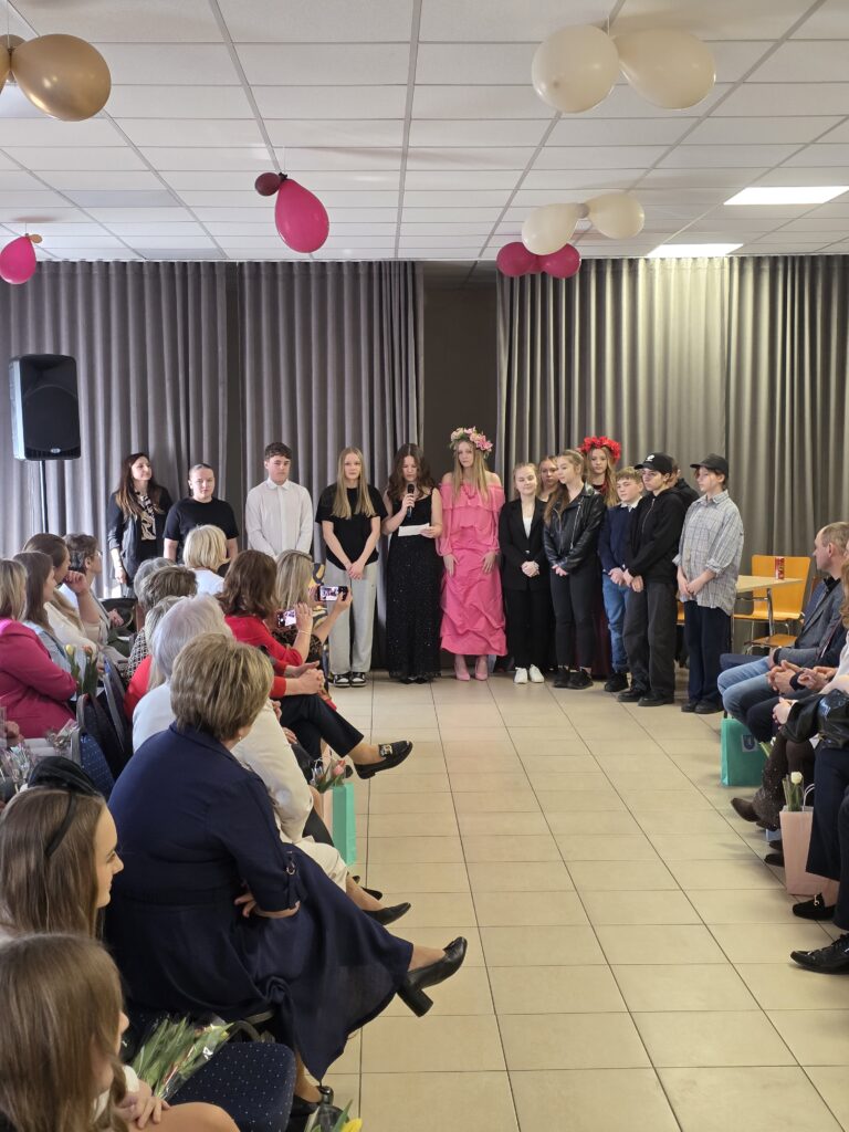 Grupa młodych ludzi stoi na scenie, niektórzy mają kolorowe kwiaty we włosach. Pomieszczenie zdobią różowe i beżowe balony. Widownia siedzi z obu stron podłogi wyłożonej płytkami.