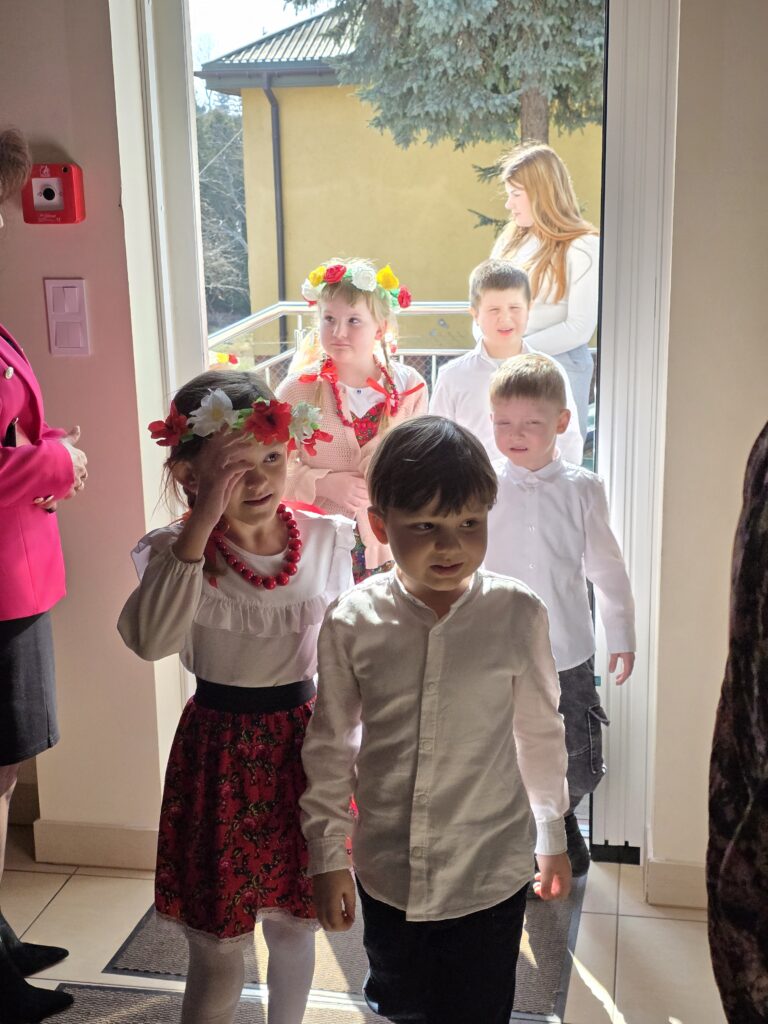 Na zdjęciu jest grupa dzieci ubranych odświętnie, wchodzących do budynku. Tło to żółty budynek oraz drzewo. Dzieci mają na sobie białe koszule i tradycyjne elementy, w tym kwiatowe wianki.