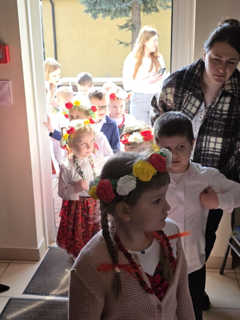 Grupa dzieci w ludowych strojach z kwiecistymi wiankami wchodzi do pomieszczenia. Ciepłe światło, kremowe ściany. Tło zewnętrzne z drzewem i żółtą elewacją.