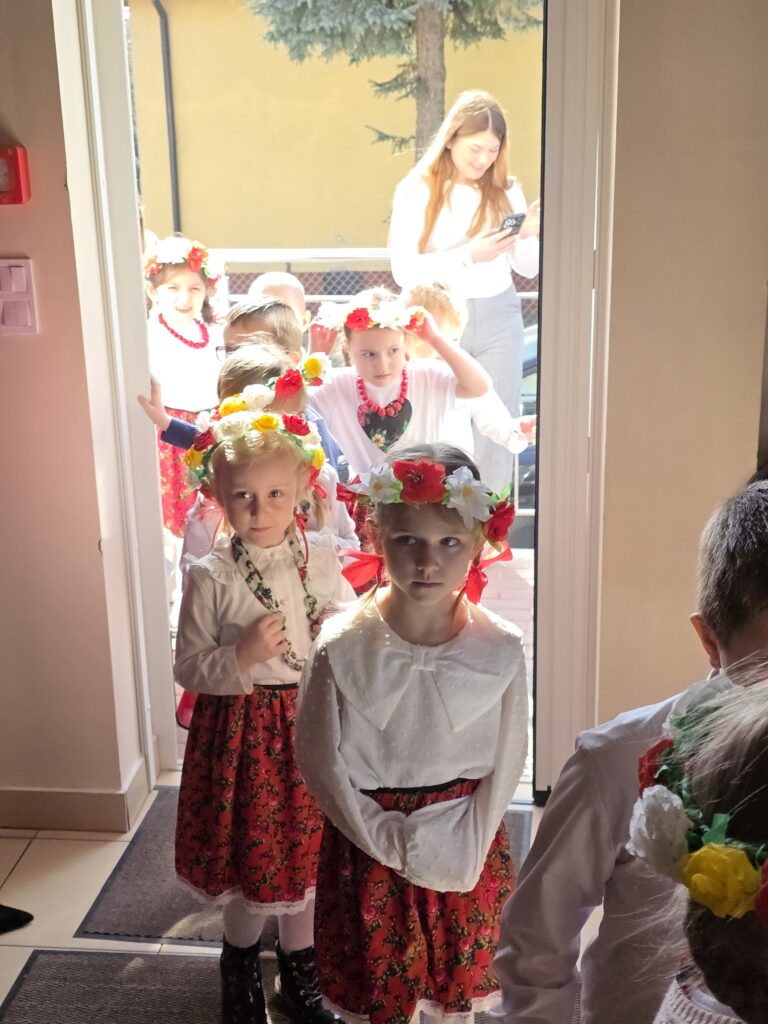 Dzieci w strojach ludowych z wiankami na głowach stoją w kolejce. Jasne wnętrze, w tle kobieta z telefonem. Za drzwiami widać żółtą ścianę i drzewo.