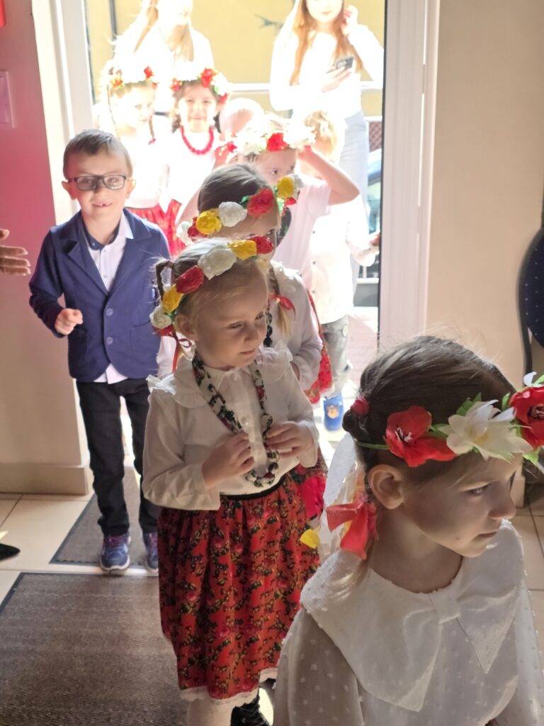 Grupa dzieci w ludowych strojach wchodzi do budynku. Mają kolorowe wianki na głowach i białe koszule. Tło jest jasne z drzwiami.