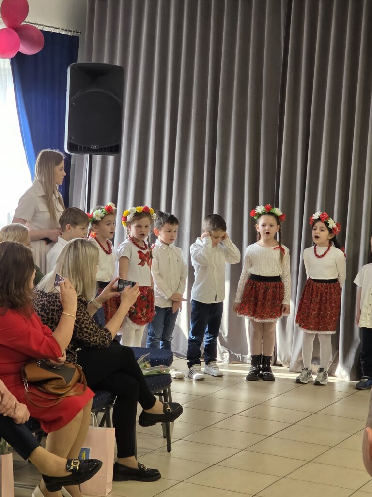 Grupa dzieci w tradycyjnych strojach wykonuje występ. W tle szare zasłony, po lewej głośnik i osoby robiące zdjęcia. Dzieci mają kolorowe wianki i spódniczki.