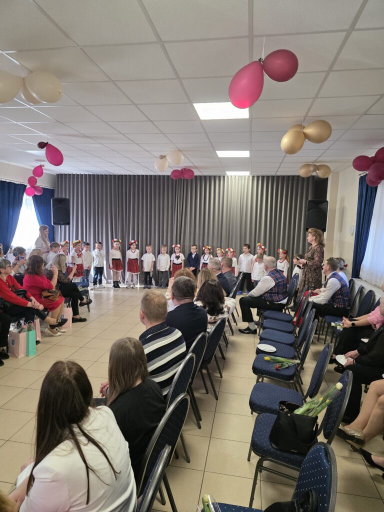 Grupa dzieci w strojach ludowych występuje na scenie. Sala jest udekorowana balonami w kolorach różowym i beżowym. Publiczność siedzi na krzesłach, a tło stanowią szare zasłony.