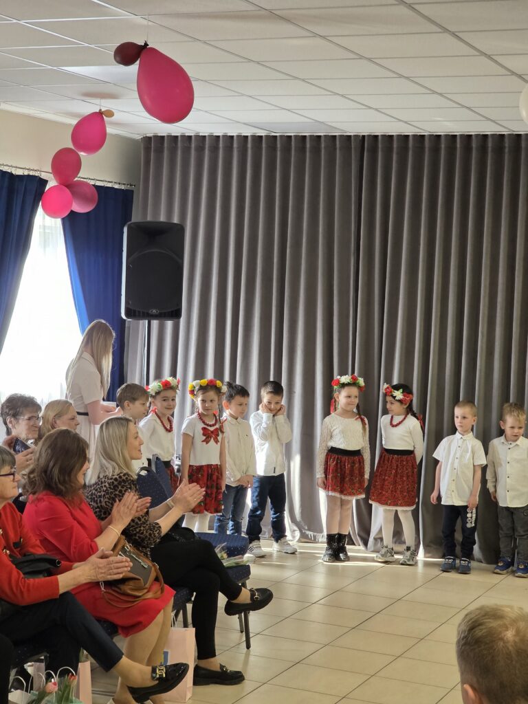 Na zdjęciu widać grupę dzieci w strojach ludowych stojących przed publicznością. W tle są szare zasłony i różowe balony pod sufitem. Dorośli siedzą i klaszczą, tworząc radosną atmosferę.