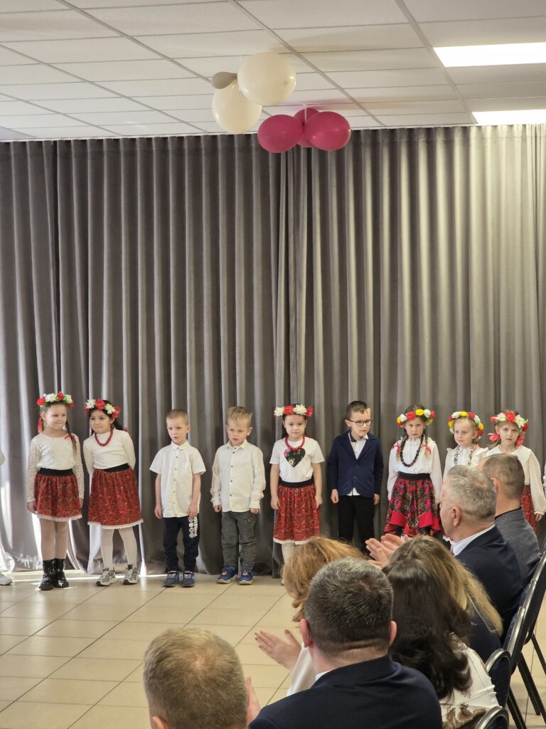 Dzieci w strojach ludowych stoją na tle szarej zasłony. Dziewczynki mają wianki, chłopcy białe koszule. Na suficie wiszą różowe i białe balony.