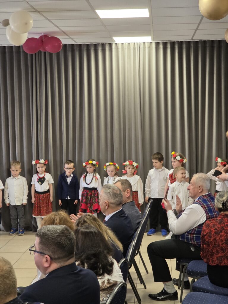 Grupa dzieci w strojach ludowych na scenie, tło z szarymi zasłonami. Dzieci mają kolorowe wieńce. Widownia siedzi na niebieskich krzesłach.