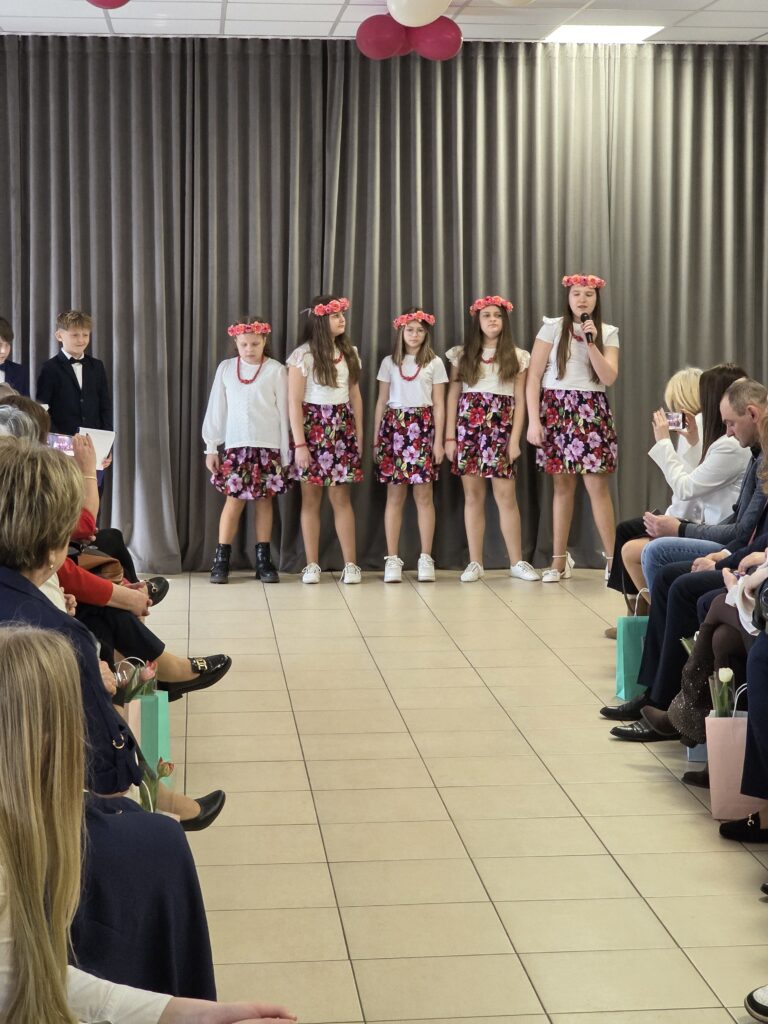 Grupa dzieci stoi na scenie przed zasłoną, ubrane w białe bluzki i kwieciste spódnice. Dziewczynki mają kwiatowe wianki na głowach. Publiczność siedzi z obu stron sali.