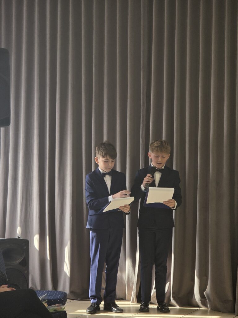 Dwóch chłopców w eleganckich garniturach, jeden trzyma mikrofon. Tło to zasłony w szaro-beżowym kolorze. Stoją na scenie z jasnym oświetleniem.
