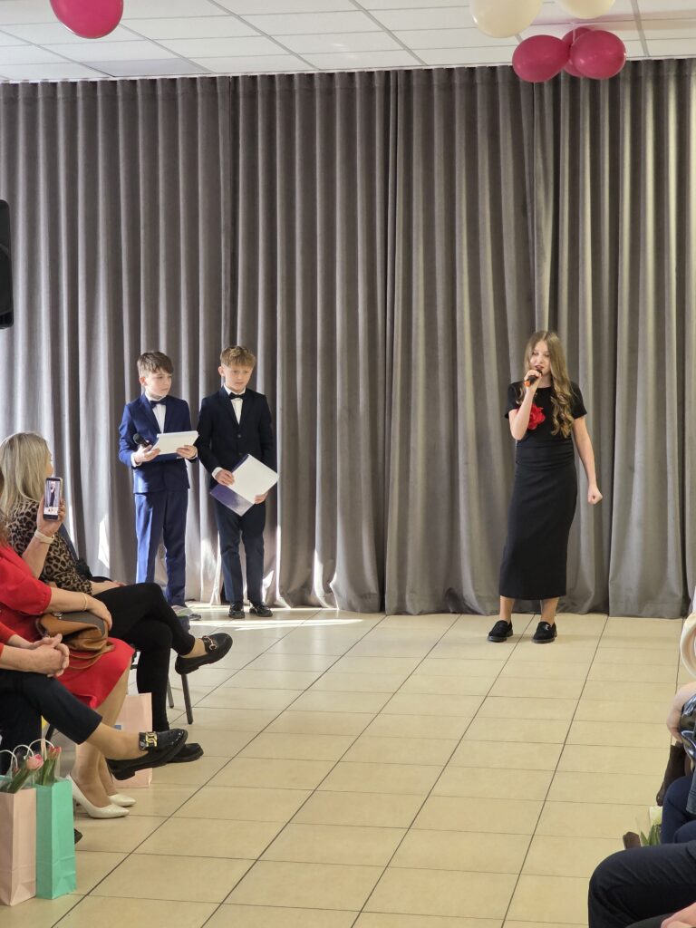 Na zdjęciu troje dzieci na scenie: dwóch chłopców w garniturach z kartkami i dziewczynka śpiewająca do mikrofonu. Tło stanowią szare zasłony, a nad nimi unoszą się balony. Widzowie siedzą na krzesłach z przodu.