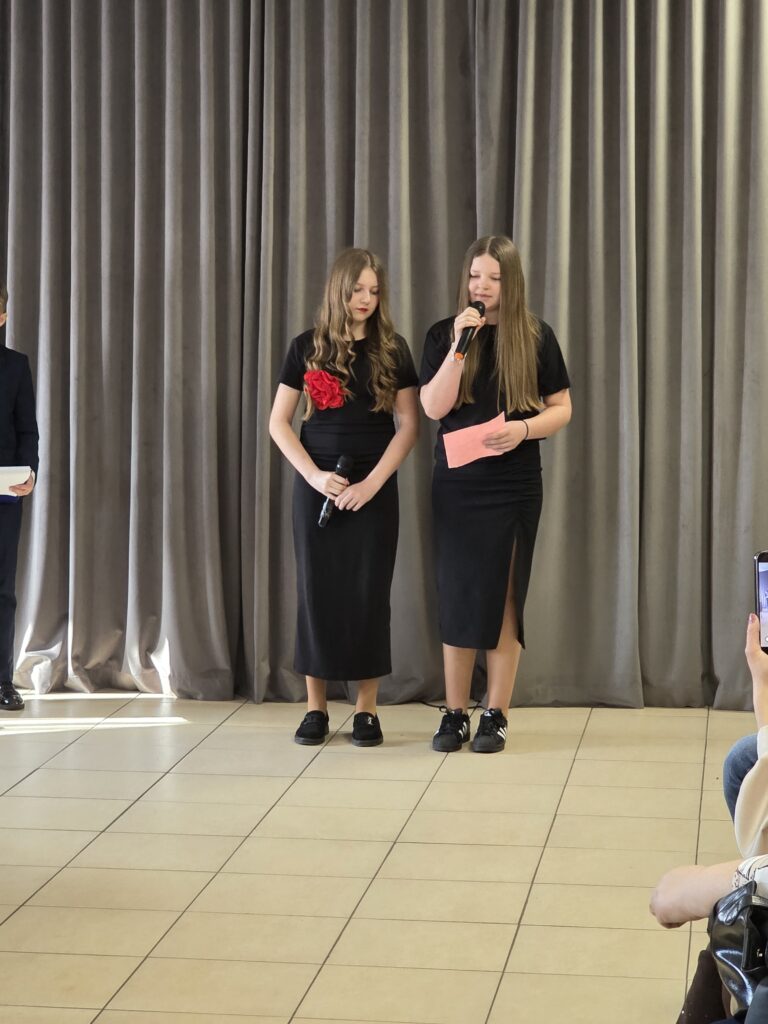 Na zdjęciu dwie dziewczyny w czarnych strojach z mikrofonami na tle szarych zasłon. Jedna ma czerwoną różę na sukience. Podłoga jest jasno beżowa.