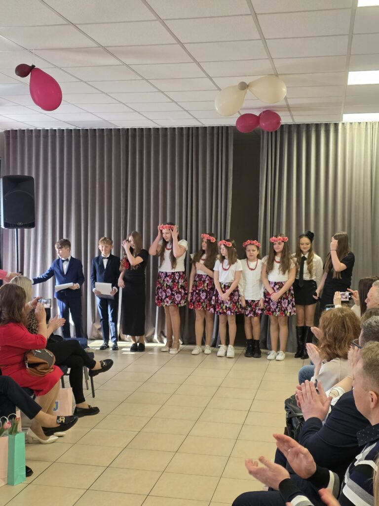 Dzieci w odświętnych strojach stoją w rzędzie na tle zasłon. Na suficie unoszą się różowe i białe balony. Widownia siedzi i klaska w jasnej sali.