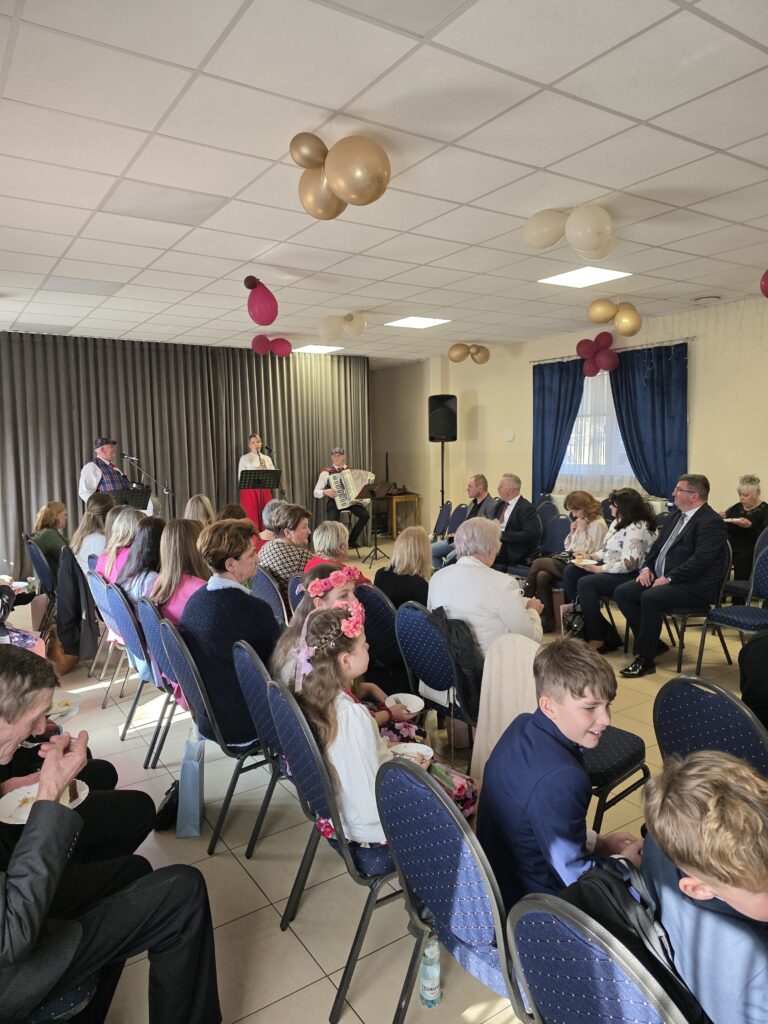 Na zdjęciu widoczne jest zgromadzenie w sali z dużymi, niebieskimi zasłonami. Ludzie siedzą na krzesłach, a z przodu trójka muzyków gra na scenie. Sufit zdobią złote i różowe balony.