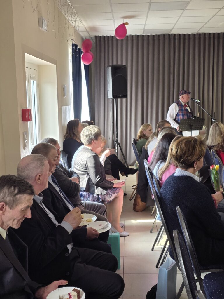 Grupa osób siedzi w jasnej sali z szarymi zasłonami. Mężczyzna przy mikrofonie, różowe balony na ścianach. Ludzie jedzą ciasto na białych talerzykach.