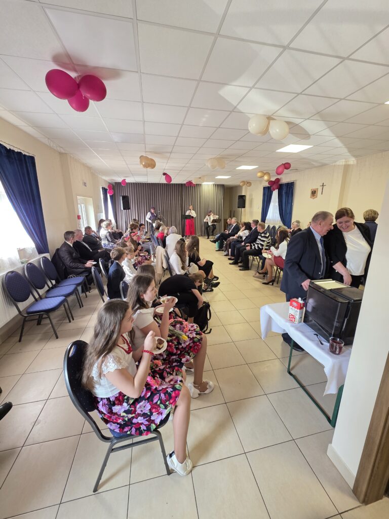 Sala z ludźmi siedzącymi na krzesłach wokół, jasne beżowe płytki na podłodze. Sufit zdobią różowe i beżowe balony. W tle karnisze zasłon i osoby przemawiające do mikrofonu.