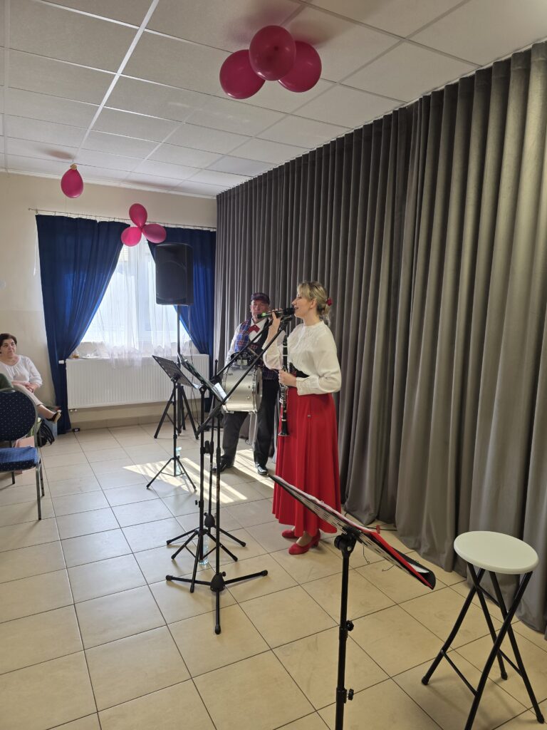 Na zdjęciu kobieta w białej bluzce i czerwonej spódnicy śpiewa do mikrofonu w jasnym pomieszczeniu. W tle szare zasłony, a z sufitu zwisają różowe balony. Obok niej stoi mężczyzna w kapeluszu przy staty