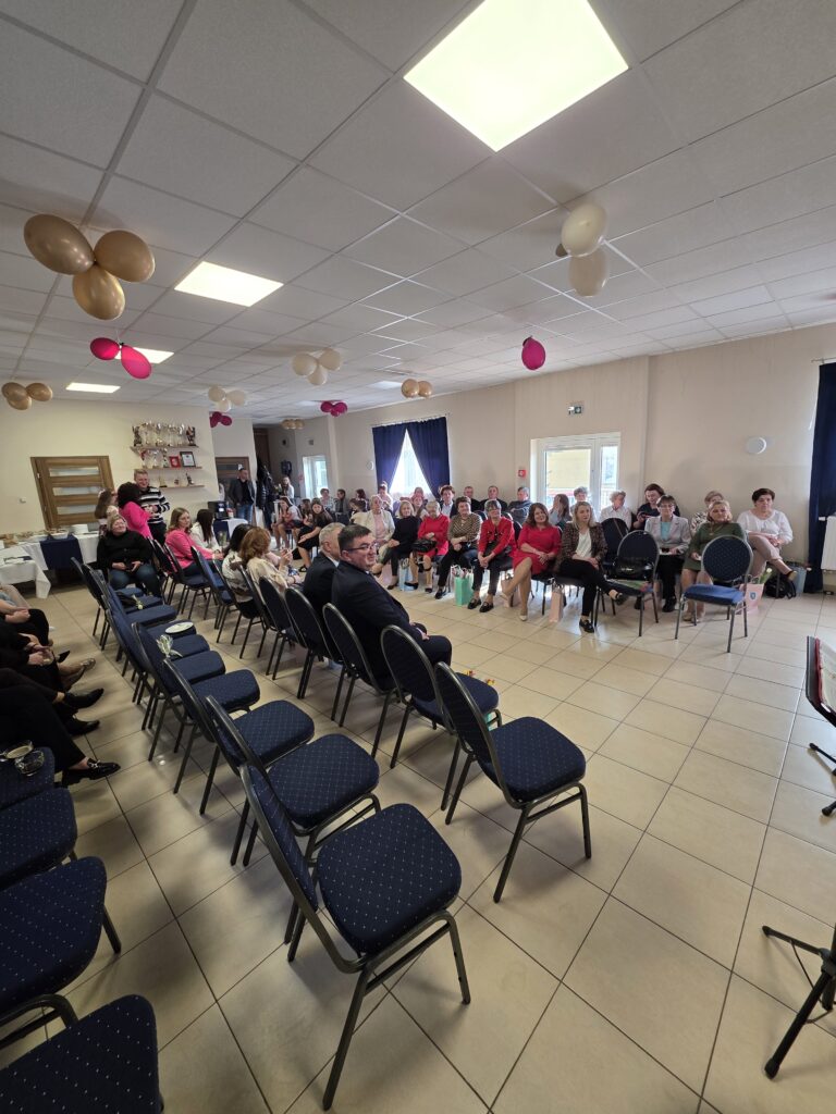 Sala z rzędem niebieskich krzeseł, w tle siedzące osoby. Na suficie wiszą złote i różowe balony. Ściany w jasnym kolorze, dekoracje dodają świątecznego nastroju.