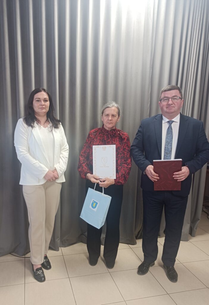 Na zdjęciu trzy osoby stoją przed szarymi zasłonami. Kobieta w środku trzyma dokument i niebieską torbę. Są elegancko ubrani, tło jest proste.