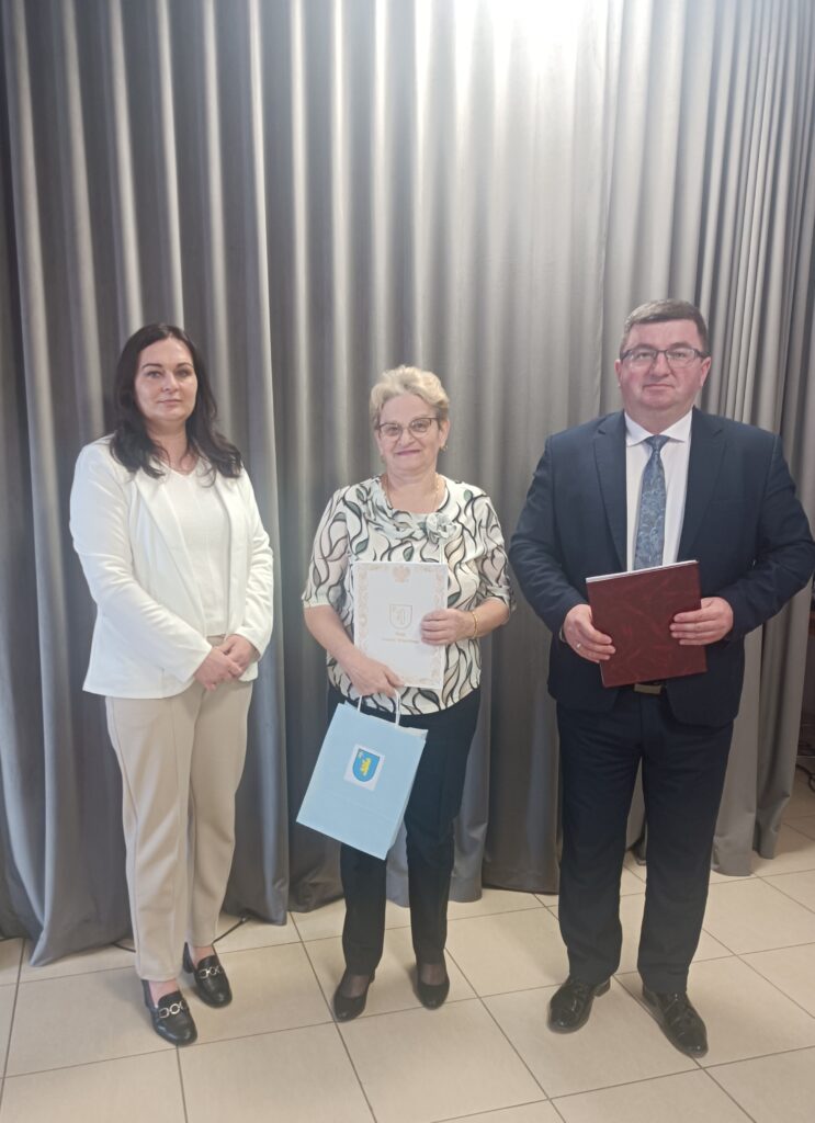 Na zdjęciu troje ludzi stoi przed szarymi zasłonami. Środkowa osoba trzyma niebieską torbę i dokument. Pozostali ubrani elegancko.