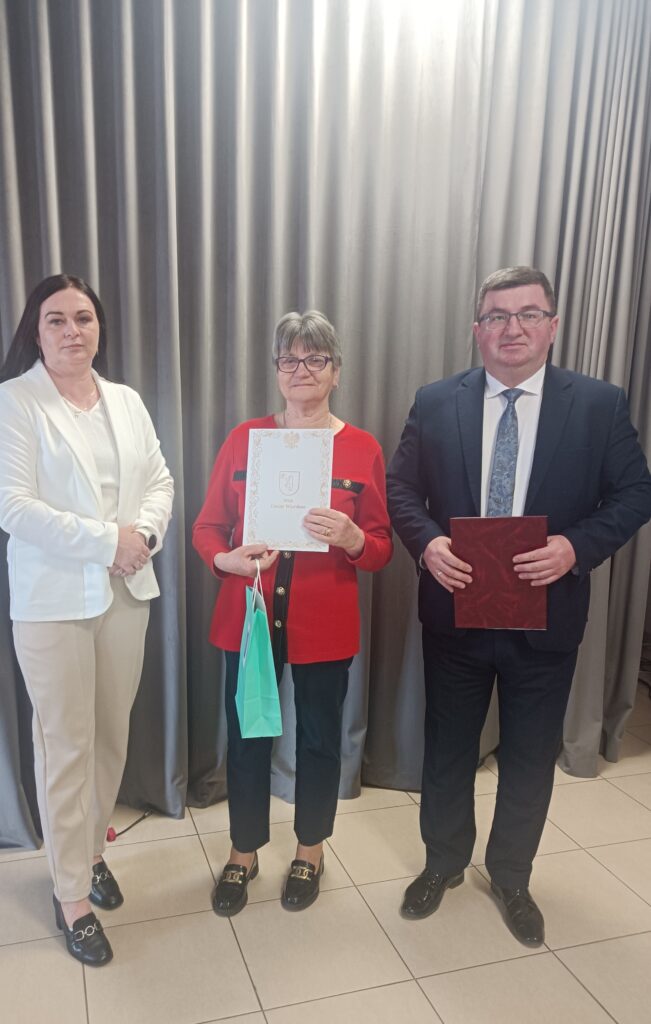 Trzy osoby stoją przed szarymi zasłonami. Środkowa osoba trzyma dokument i torbę. Wszyscy są elegancko ubrani.