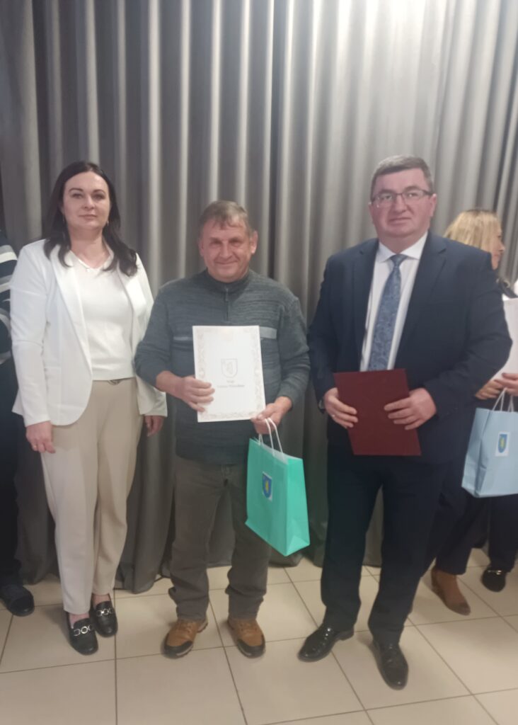 Trzy osoby stoją razem na tle szarych zasłon. Osoba pośrodku trzyma certyfikat i niebieską torbę. Ubrania w odcieniach bieli, szarości i ciemnych kolorach.