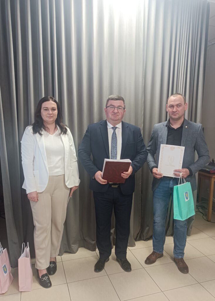 Trzy osoby stoją na tle szarej zasłony. Środkowa w garniturze, trzyma teczkę. Po bokach osoby trzymają torby.