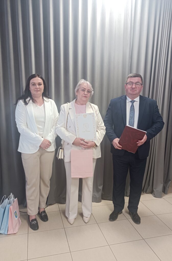 Trzy osoby stoją na tle szarej zasłony. Dwie kobiety noszą białe ubrania. Mężczyzna ubrany jest w ciemny garnitur, trzyma teczkę.