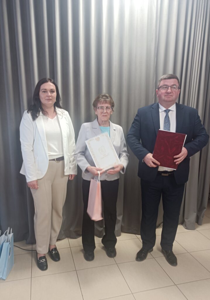 Trzy osoby stoją na tle szarych zasłon. Środkowa osoba trzyma dyplom, a reszta książki. Ubrania są eleganckie i stonowane.