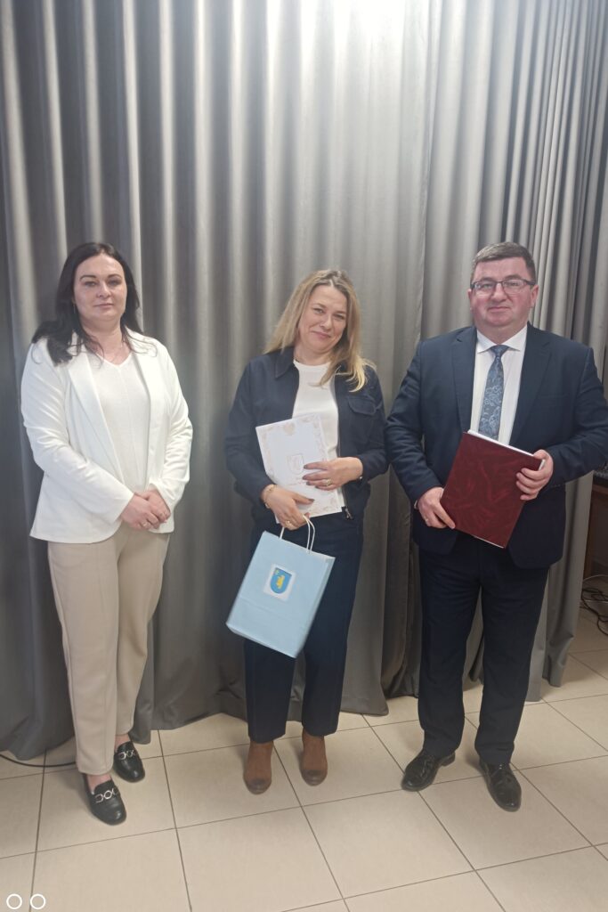 Trzy osoby stoją na tle szarych zasłon. Każda z nich trzyma dokumenty. Osoby ubrane są elegancko.