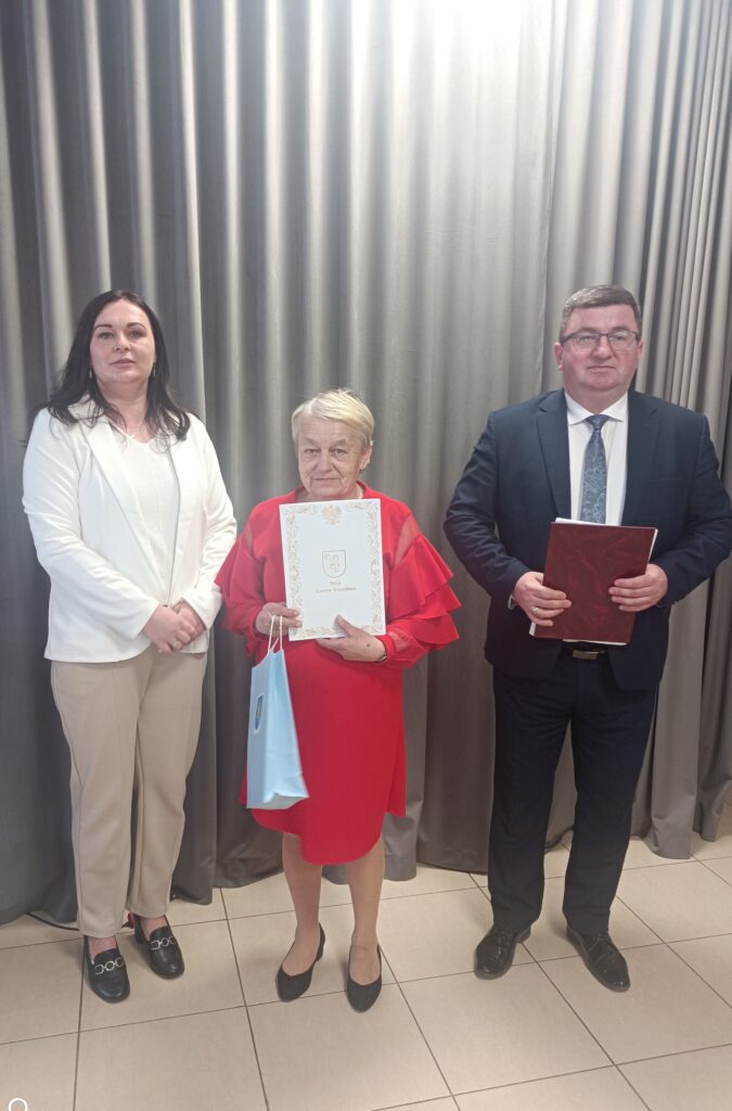 Na zdjęciu trzy osoby w eleganckich strojach stoją na tle szarej zasłony. Kobieta w czerwieni trzyma dyplom. Płytki podłogowe są jasne.