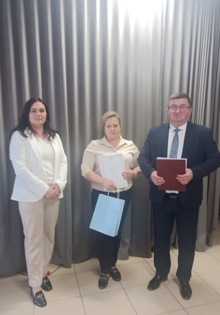 Trzy osoby stoją na tle szarych zasłon. Kobieta po lewej w białym stroju, środkowa trzyma niebieską torebkę i dokument. Mężczyzna po prawej w garniturze trzyma czerwoną teczkę.