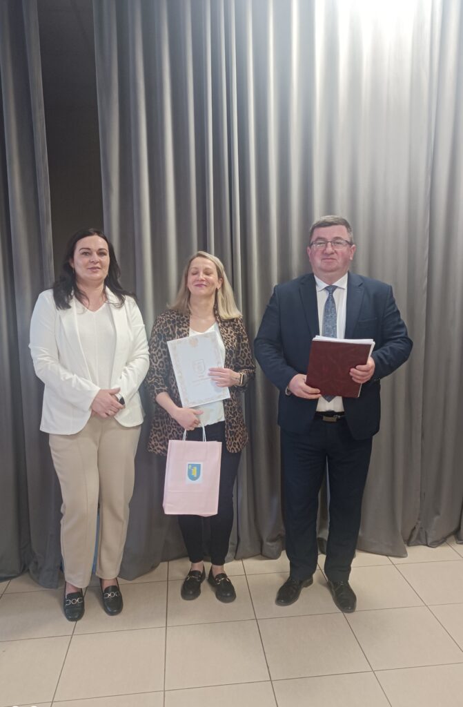 Trzy osoby stoją na tle szarych zasłon. Kobieta pośrodku trzyma dyplom i różową torbę. Wszyscy są elegancko ubrani.