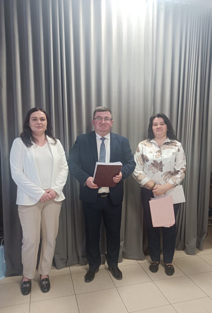 Troje osób stoi przed szarymi zasłonami. Środkowa osoba trzyma dokumenty. Lewa i prawa postać mają eleganckie ubrania.