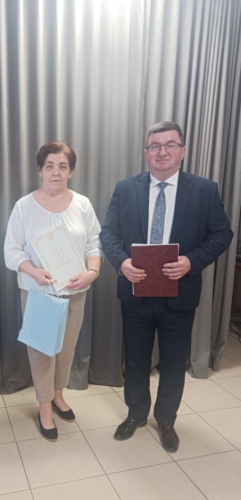 Dwie osoby w oficjalnej odzieży stoją na tle szarych zasłon. Kobieta trzyma błękitną torbę i białą teczkę, mężczyzna czerwoną teczkę. Podłoga jest wyłożona jasnymi kafelkami.